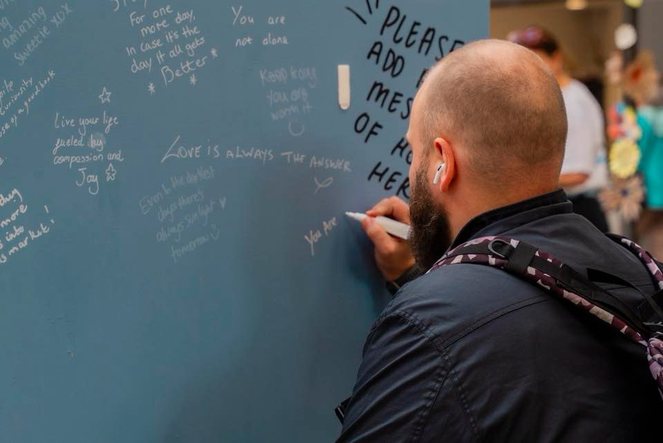 A man with a beard and headphones writes messages on a blue wall filled with inspirational quotes and affirmations.