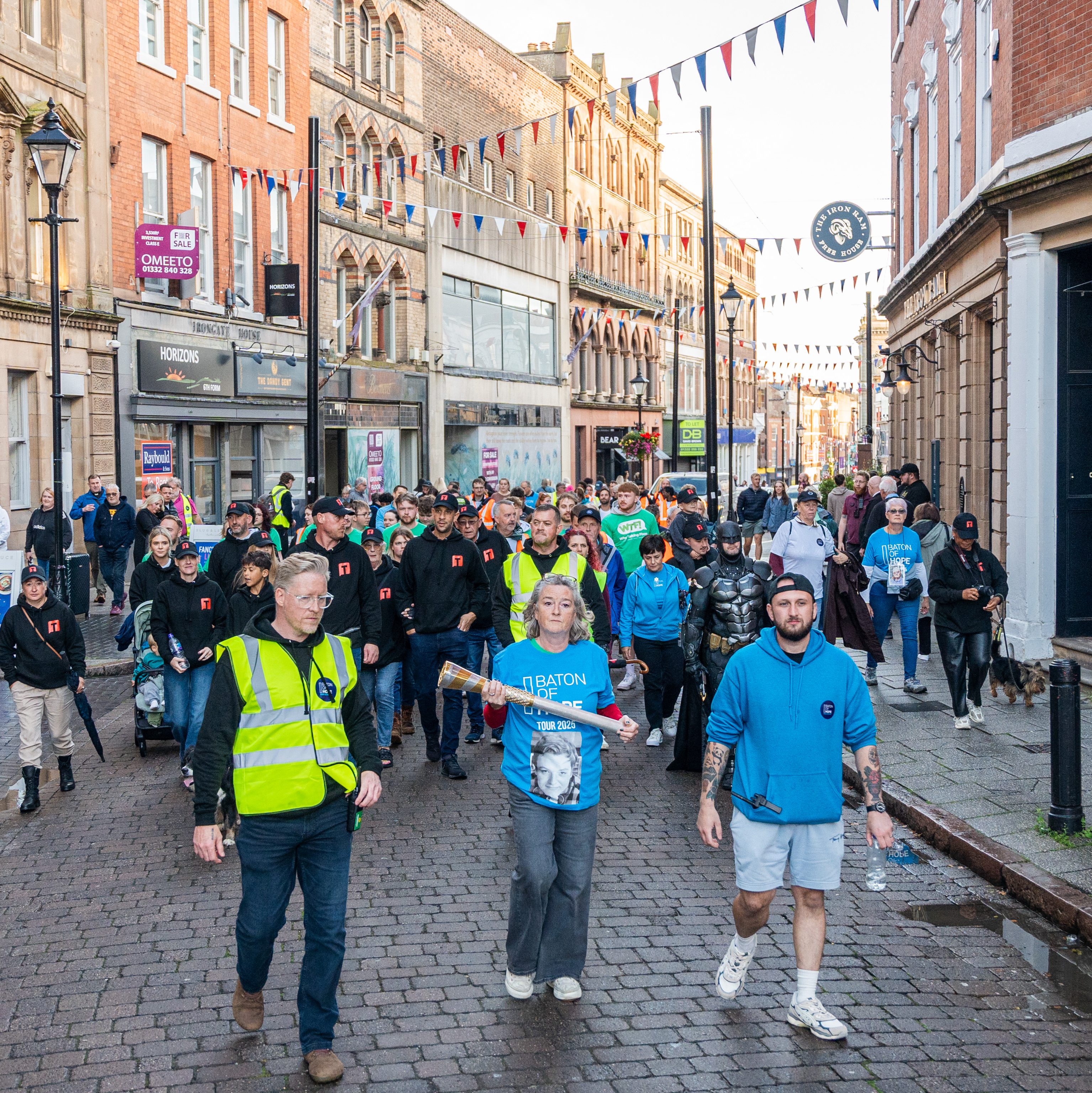 A large group of people walking down a street decorated with bunting. Some participants wear high-visibility vests, while others are in casual attire. A woman leads the group holding a baton, accompanied by a man in a blue hoodie and others in various outfits.